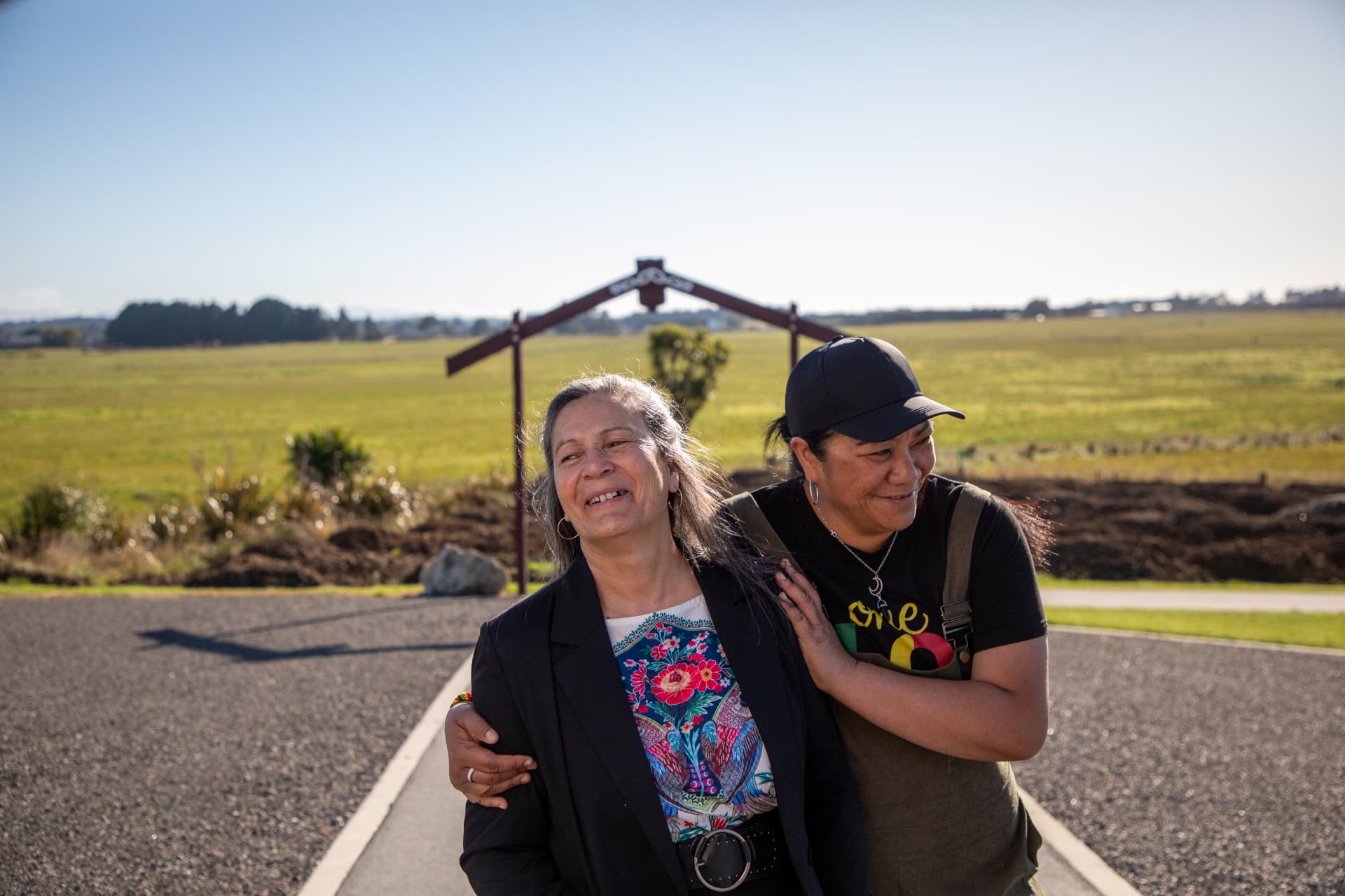 Two wāhine smiling together on the path in front of the waharoa